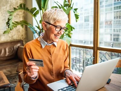 En ældre kvinde med kort hår og briller handler online på sin bærbare computer med sit bankkort i den ene hånd