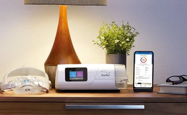 An AirCurve 11 BIPAP device, mask and mobile phone showing the myAir app on a bedside table