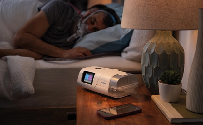 Et AirCurve 11-apparat og en mobiltelefon på et natbord, hvor brugeren af apparatet sover i sengen iført maske.